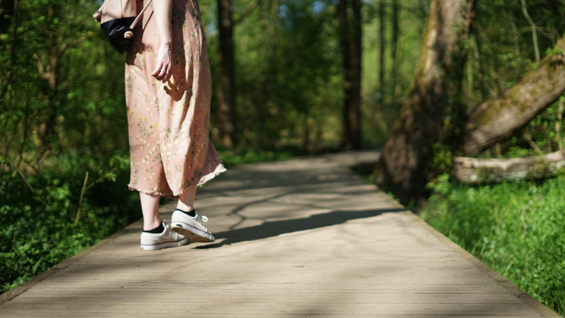 A person in a long peach dress walking away along a wooden boardwalk through woodland